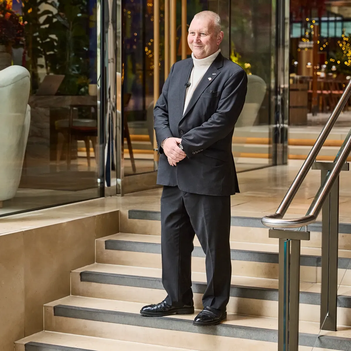 A man in a dark suit stands on steps outside a modern glass building.