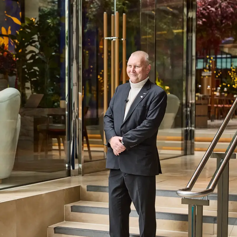 A man in a dark suit stands on steps outside a modern glass building.