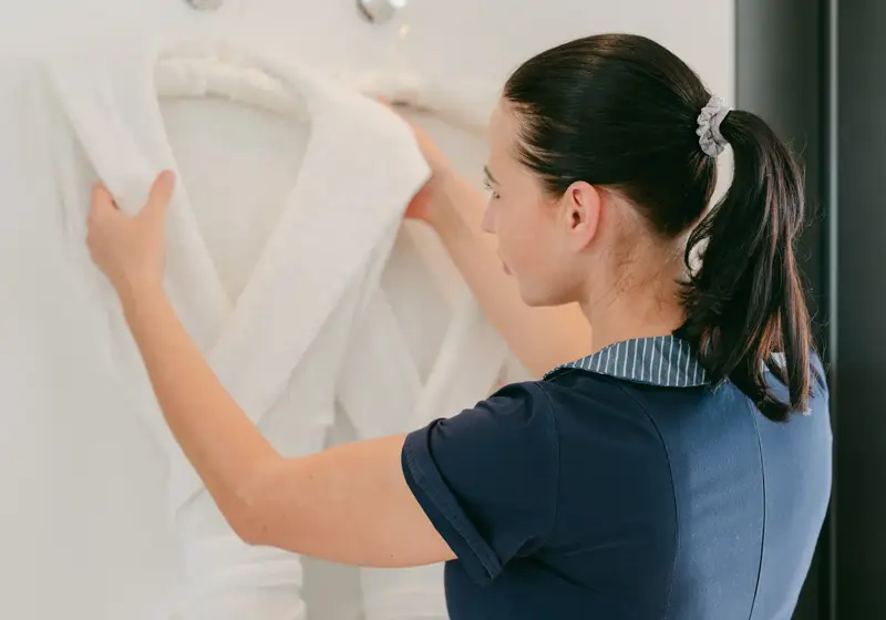 Housekeeper in a navy uniform adjusts a white bathrobe on a wall hook in a bright bathroom