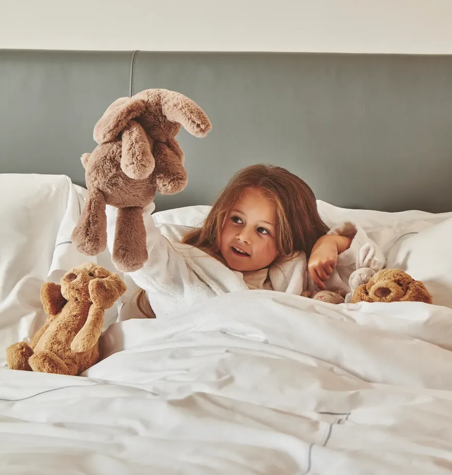 Child playing with plush toys while tucked into a white hotel bed with soft linens and upholstered headboard.