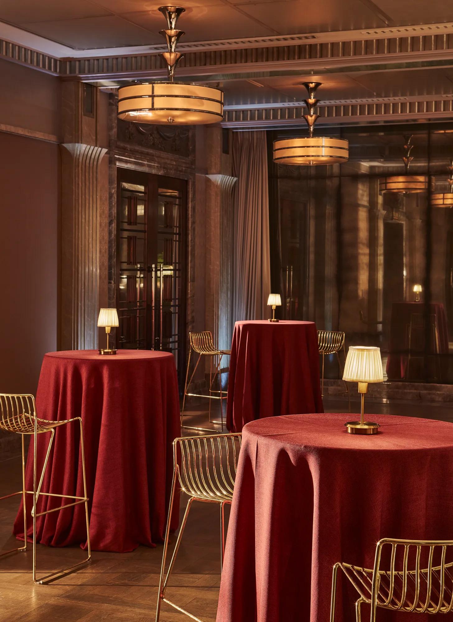 Cocktail space with tall red-draped tables, gold metal bar stools and small table lamps, set beneath warm Art Deco ceiling lights.
