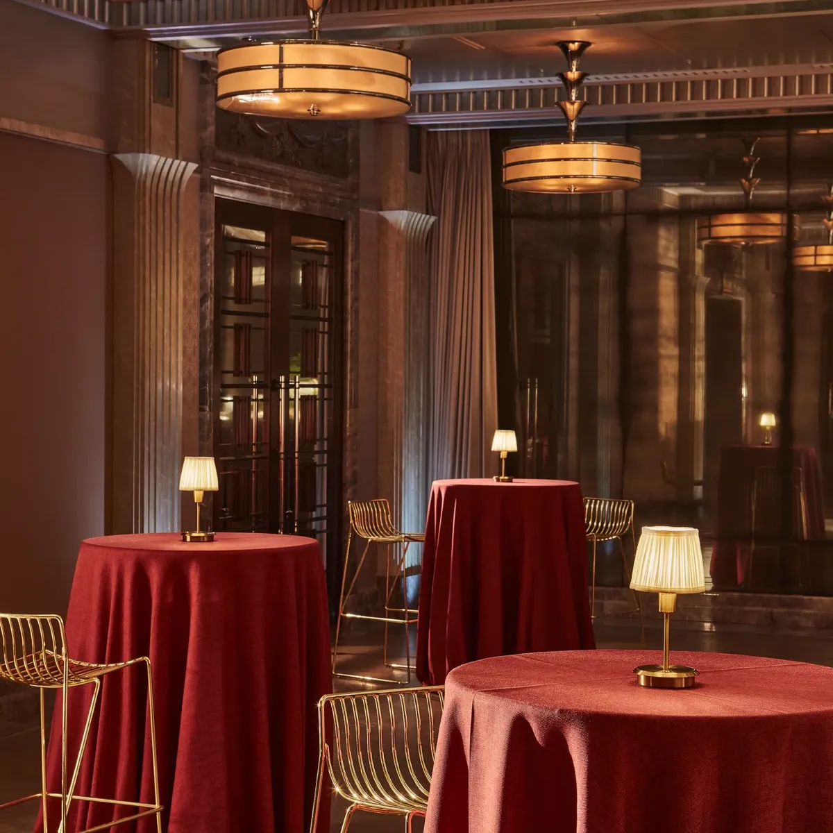 Cocktail space with tall red-draped tables, gold metal bar stools and small table lamps, set beneath warm Art Deco ceiling lights.