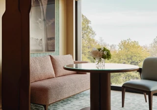 Dining nook with a round table, upholstered bench and chair, set beside a large window overlooking leafy treetops.