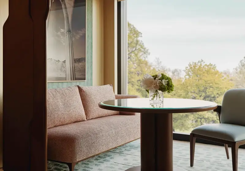 Dining nook with a round table, upholstered bench and chair, set beside a large window overlooking leafy treetops.