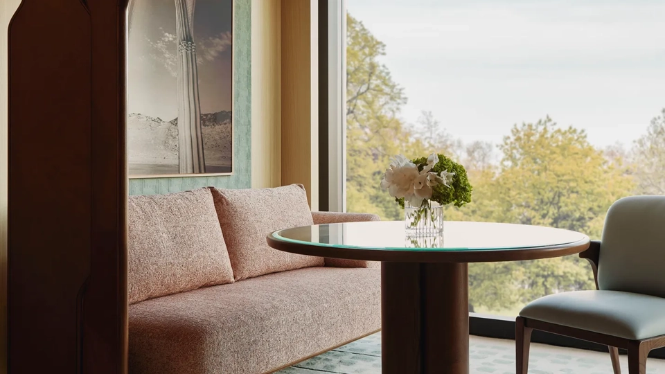 Dining nook with a round table, upholstered bench and chair, set beside a large window overlooking leafy treetops.