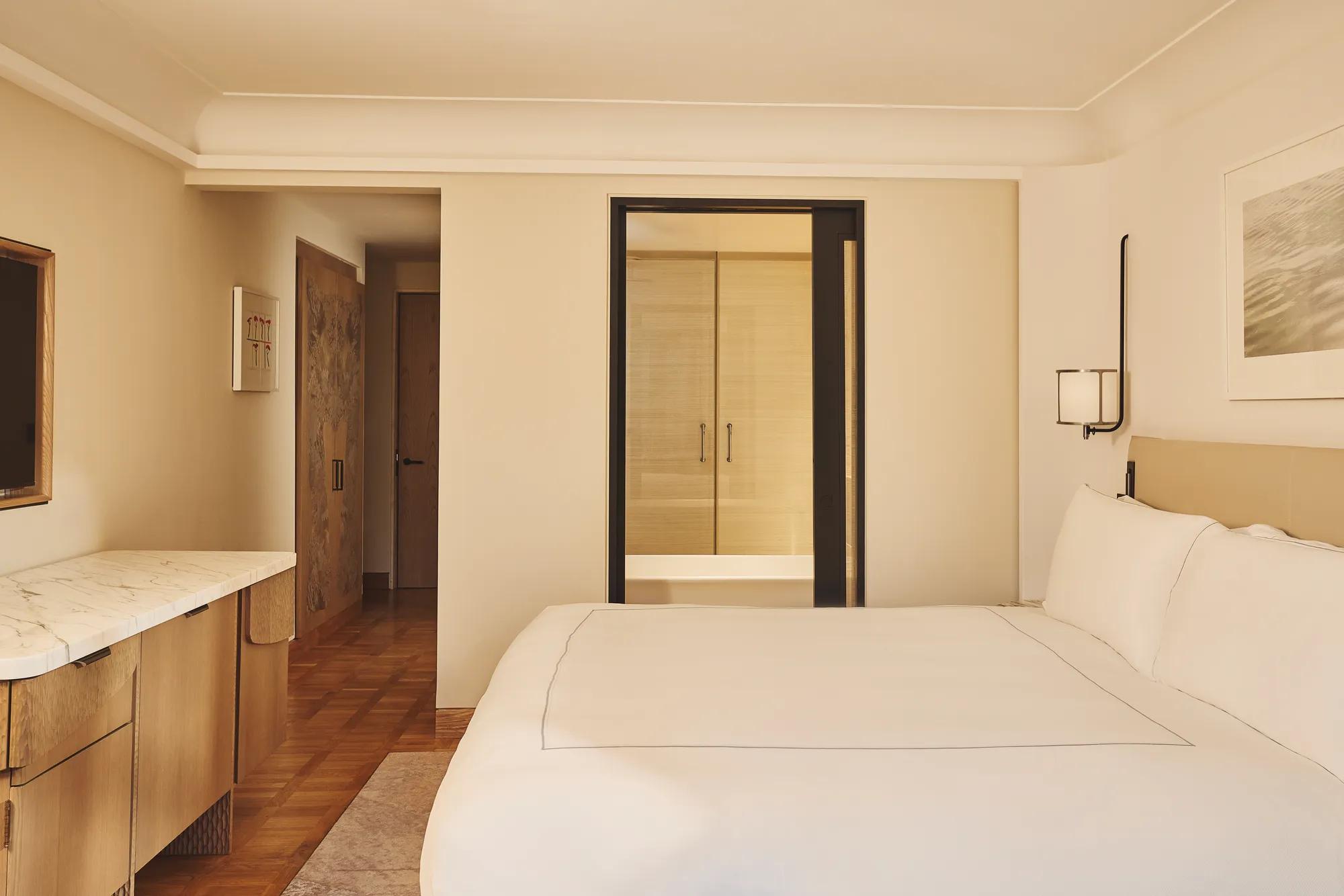 Minimal bedroom with king bed, marble desk, wood flooring, and view through to bathroom.