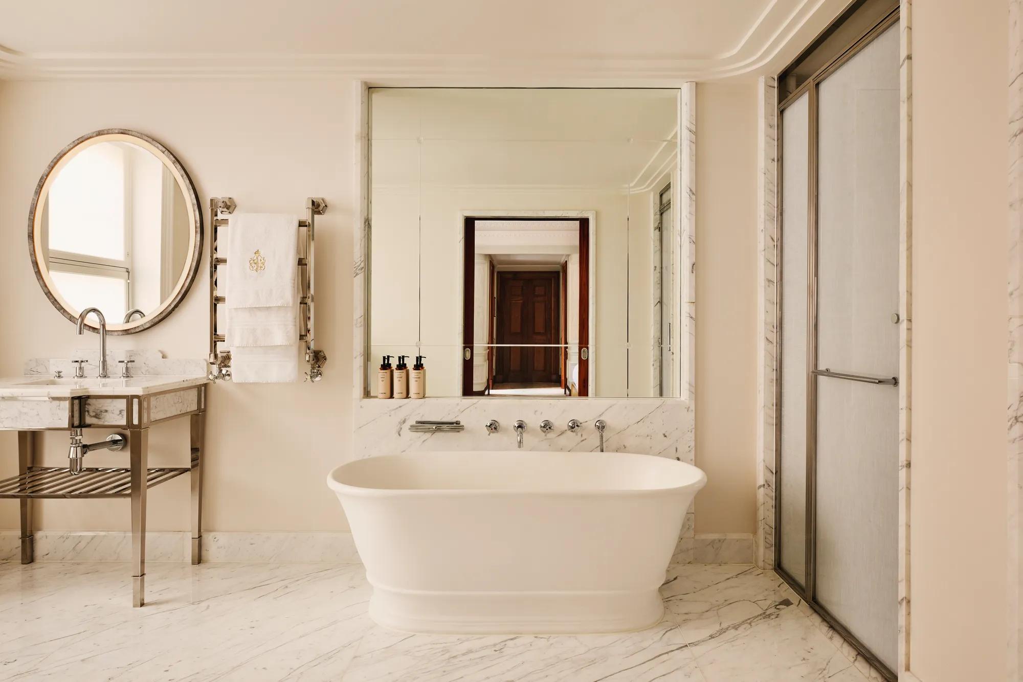 Elegant marble bathroom featuring a freestanding bathtub beneath a large mirror, a circular vanity mirror with a marble sink to the left, and a glass-enclosed shower to the right.