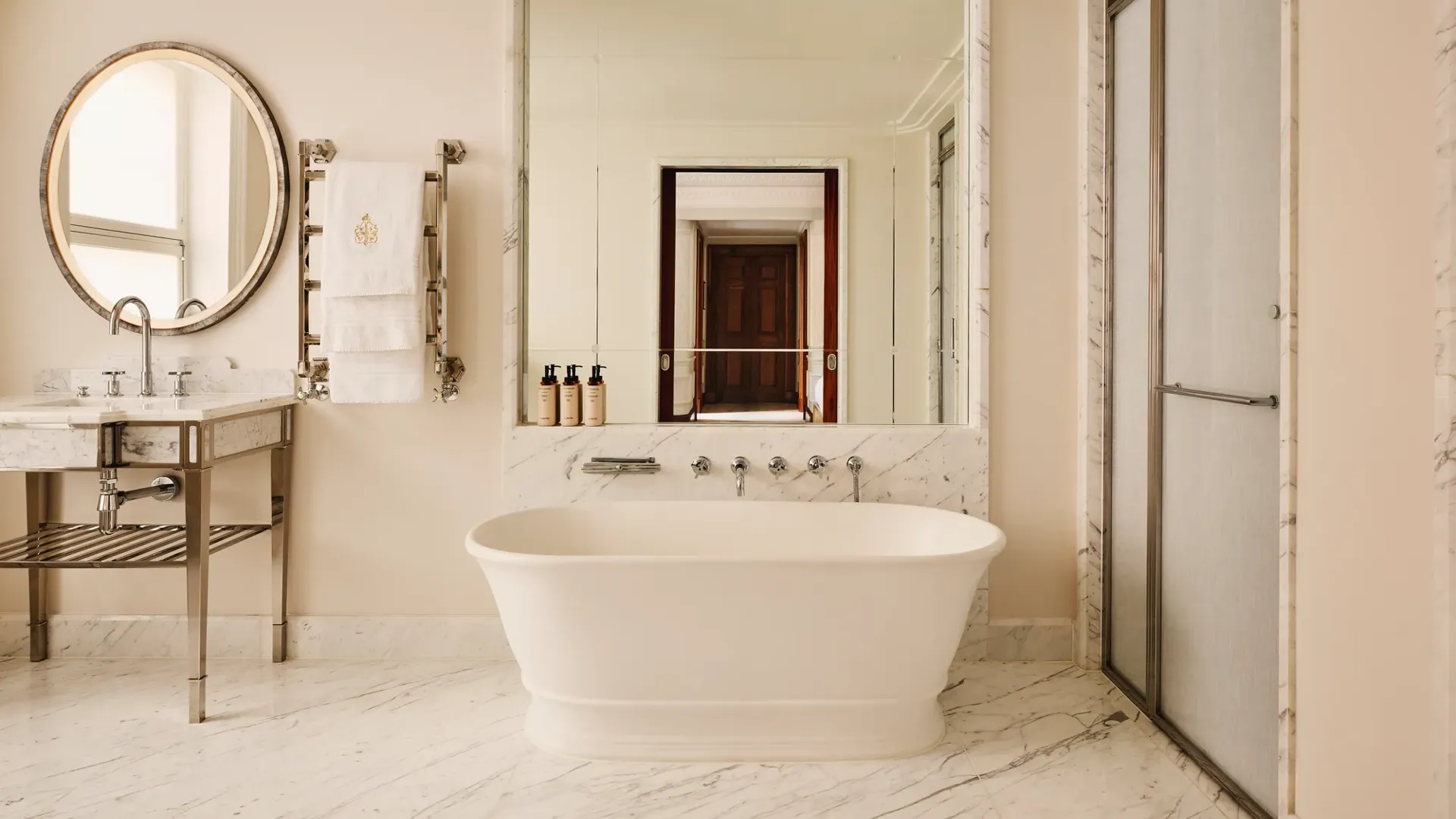 Elegant marble bathroom featuring a freestanding bathtub beneath a large mirror, a circular vanity mirror with a marble sink to the left, and a glass-enclosed shower to the right.