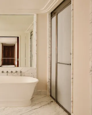 Elegant marble bathroom featuring a freestanding bathtub beneath a large mirror, a circular vanity mirror with a marble sink to the left, and a glass-enclosed shower to the right.