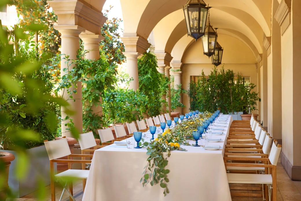 Daytime long table on the Terrace Patio loggia; blue goblets, lemons, and eucalyptus garland beneath hanging lanterns and arches.