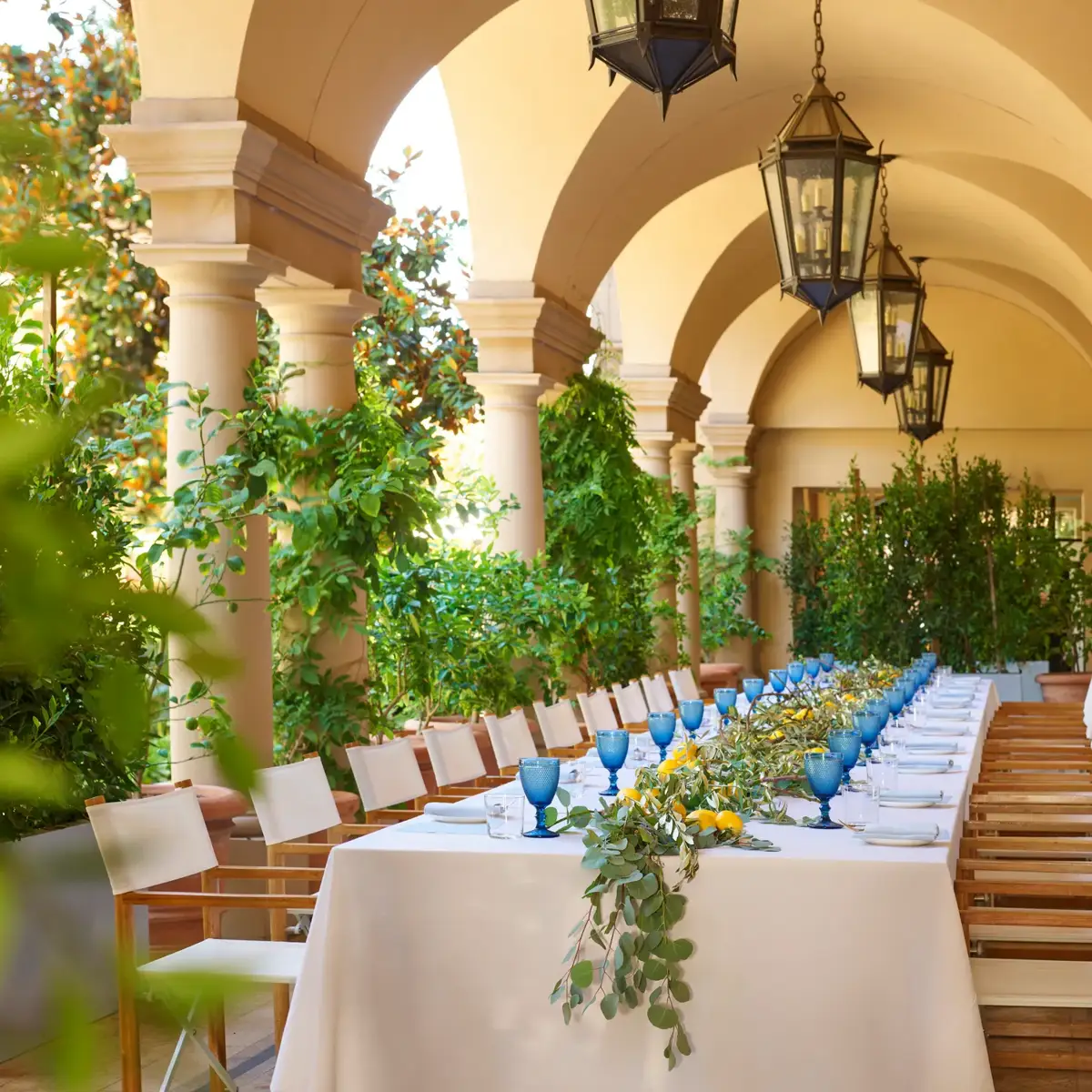 Daytime long table on the Terrace Patio loggia; blue goblets, lemons, and eucalyptus garland beneath hanging lanterns and arches.