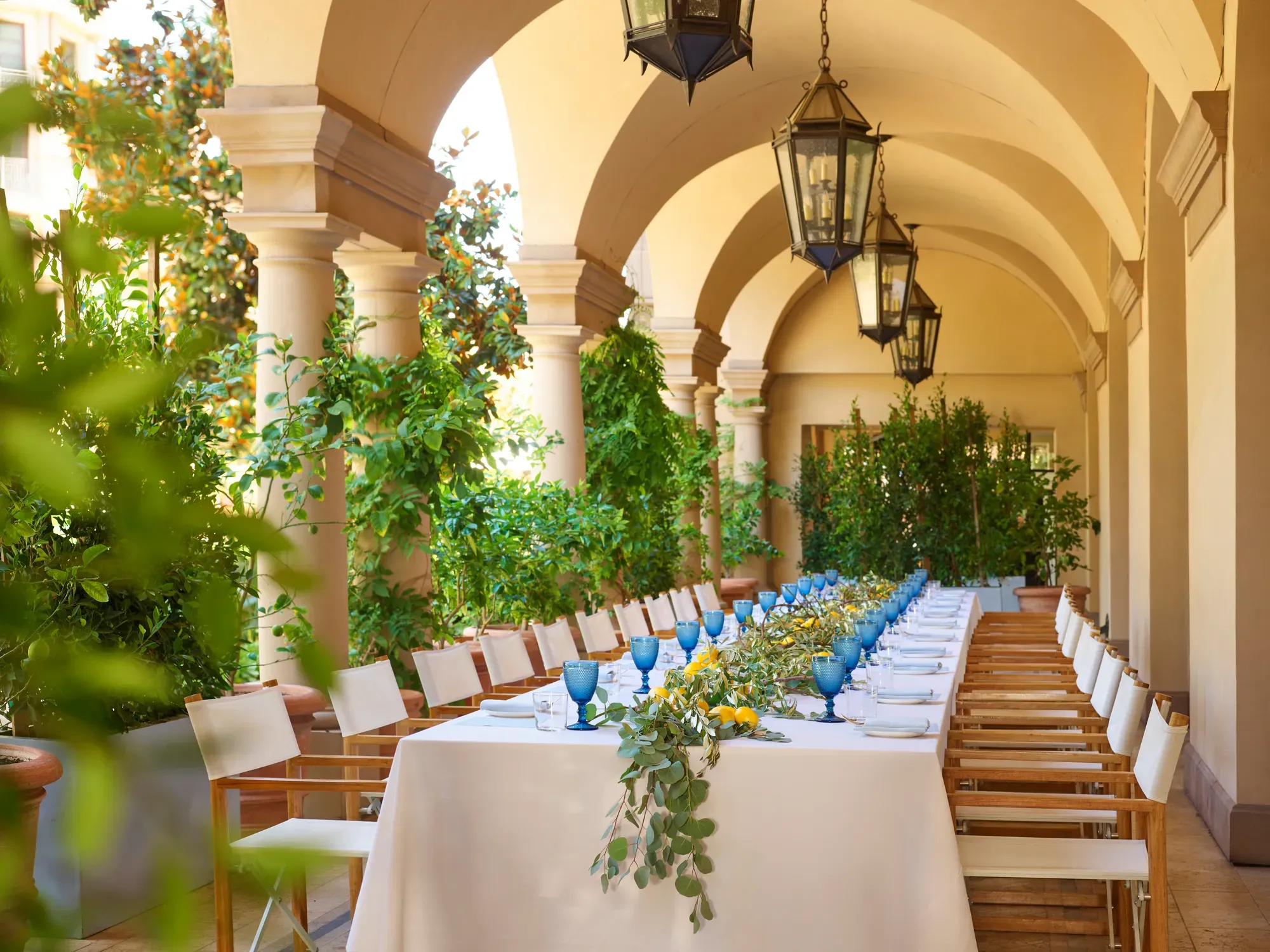 Daytime long table on the Terrace Patio loggia; blue goblets, lemons, and eucalyptus garland beneath hanging lanterns and arches.