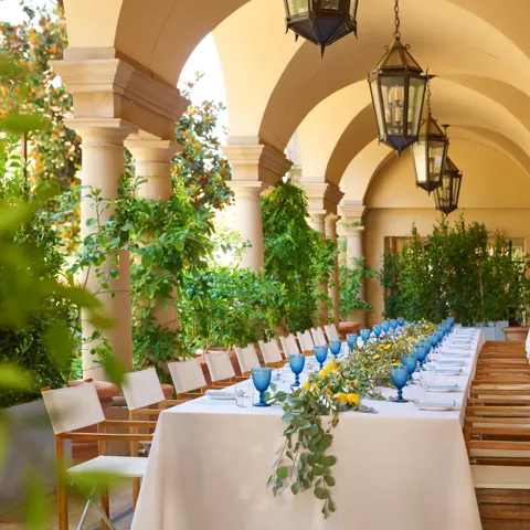 Daytime long table on the Terrace Patio loggia; blue goblets, lemons, and eucalyptus garland beneath hanging lanterns and arches.