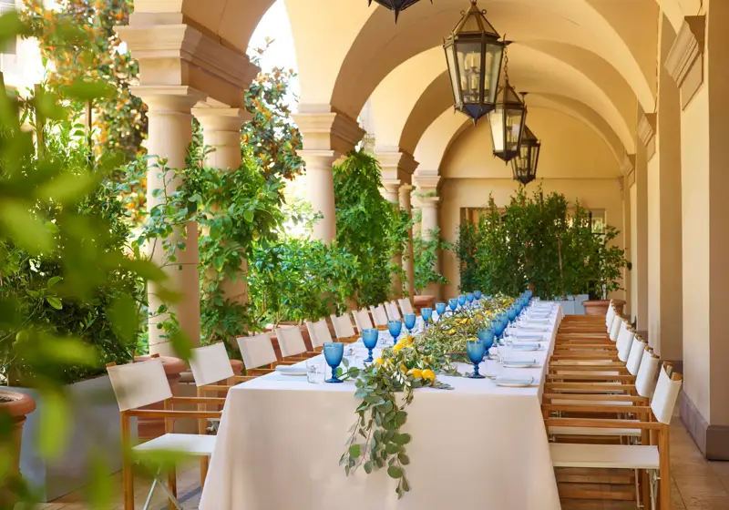 Daytime long table on the Terrace Patio loggia; blue goblets, lemons, and eucalyptus garland beneath hanging lanterns and arches.