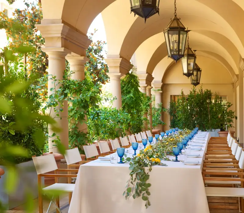 Daytime long table on the Terrace Patio loggia; blue goblets, lemons, and eucalyptus garland beneath hanging lanterns and arches.