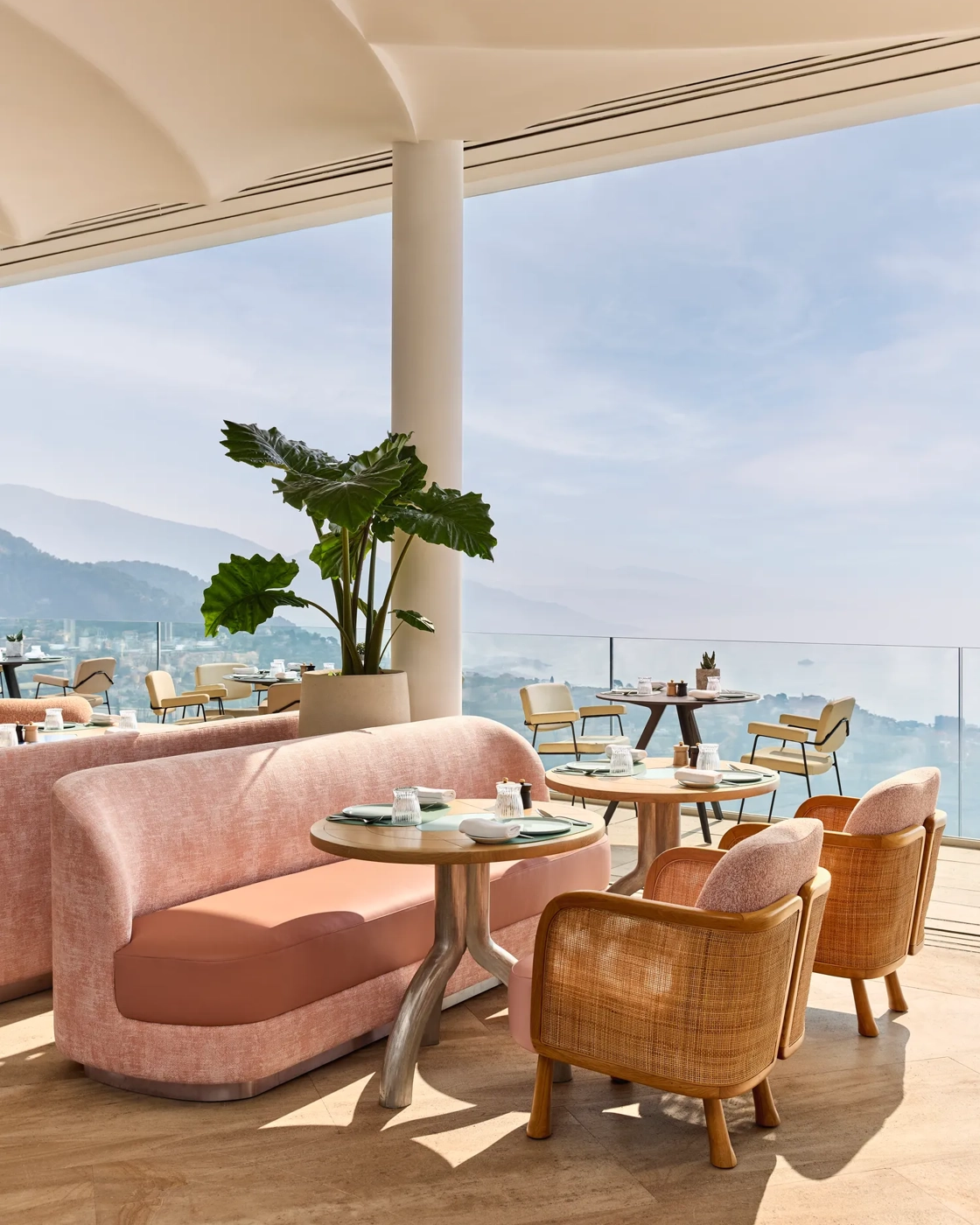 Terrasse ouverte avec des sièges pastel, des plantes en pot et une vue sur la mer sous une tonnelle crème.