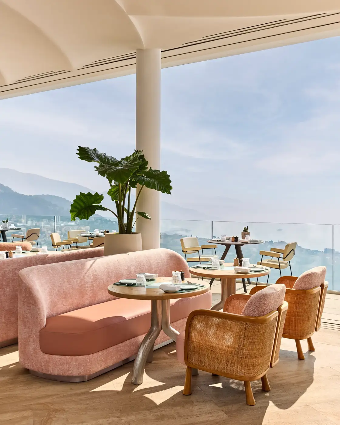 Terrasse ouverte avec des sièges pastel, des plantes en pot et une vue sur la mer sous une tonnelle crème.