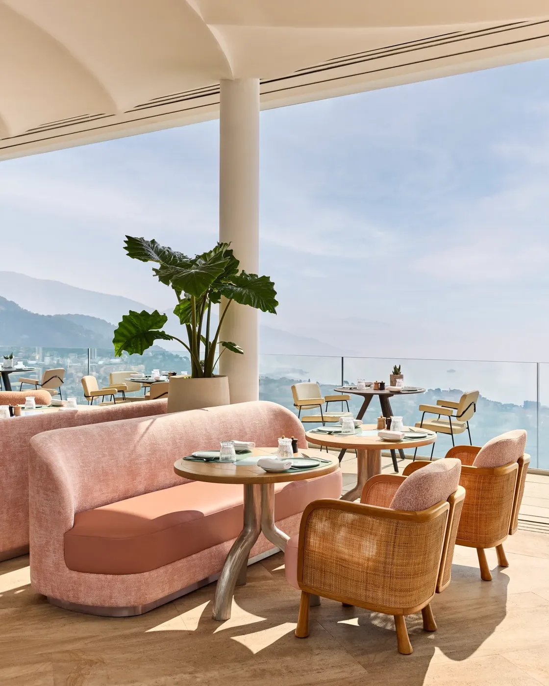Terrasse ouverte avec des sièges pastel, des plantes en pot et une vue sur la mer sous une tonnelle crème.