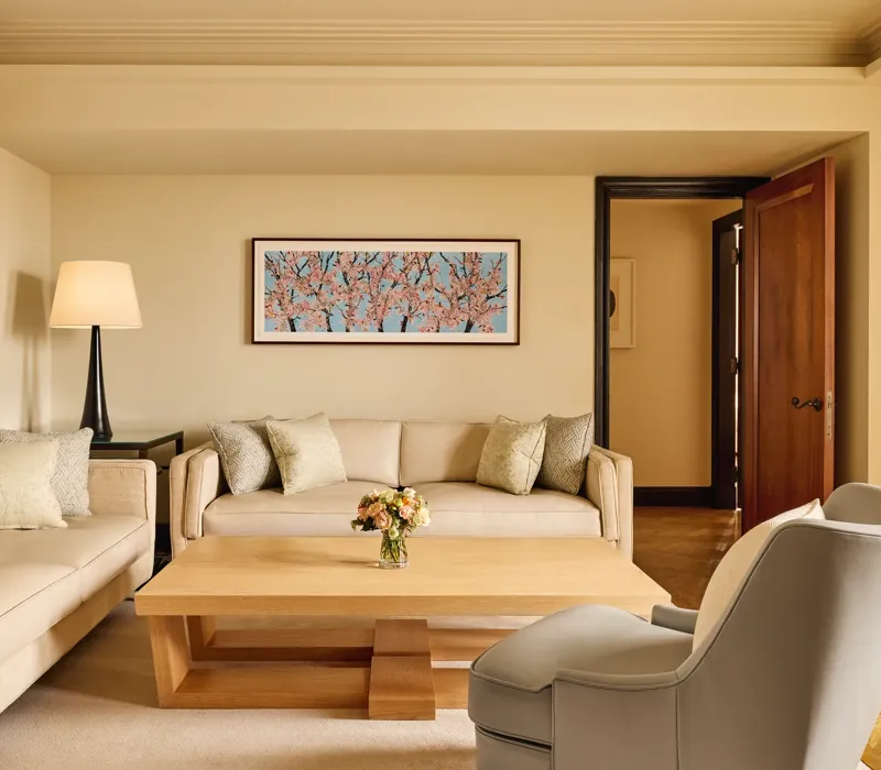 Bright living room with two cream sofas, a grey armchair, and a light wood coffee table topped with flowers, featuring a cherry blossom artwork on the wall above.