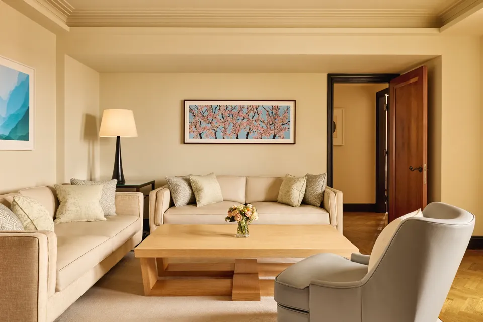 Bright living room with two cream sofas, a grey armchair, and a light wood coffee table topped with flowers, featuring a cherry blossom artwork on the wall above.