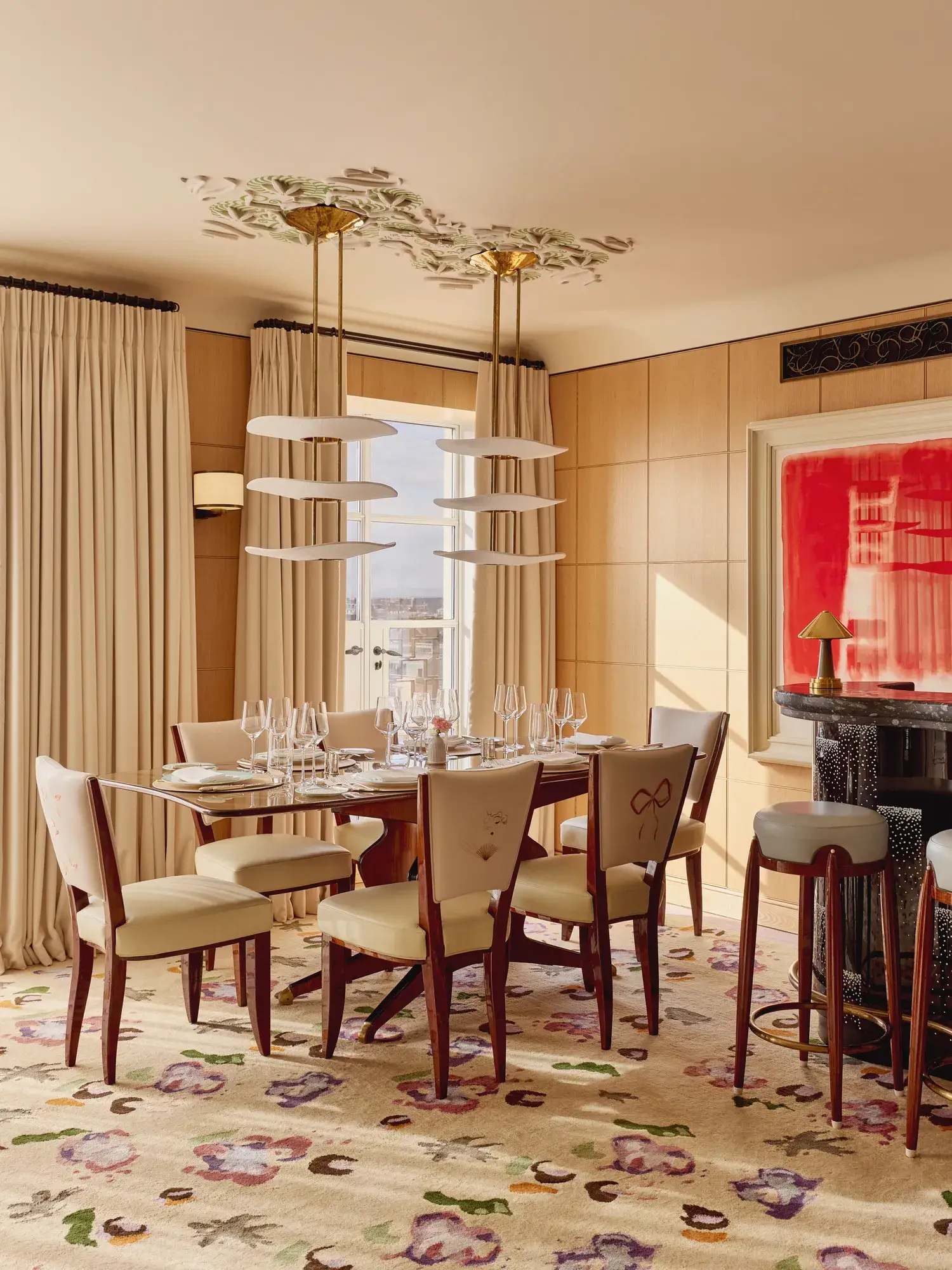 Elegant dining room with a wooden table set for six, upholstered chairs embroidered with floral motifs, a patterned rug, and a small curved bar in warm natural light.