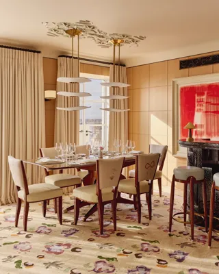 Elegant dining room with a wooden table set for six, upholstered chairs embroidered with floral motifs, a patterned rug, and a small curved bar in warm natural light.