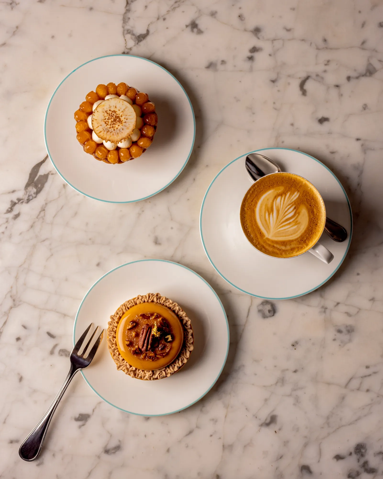 Three small pastries and a cappuccino with latte art arranged on white plates on a marble table.