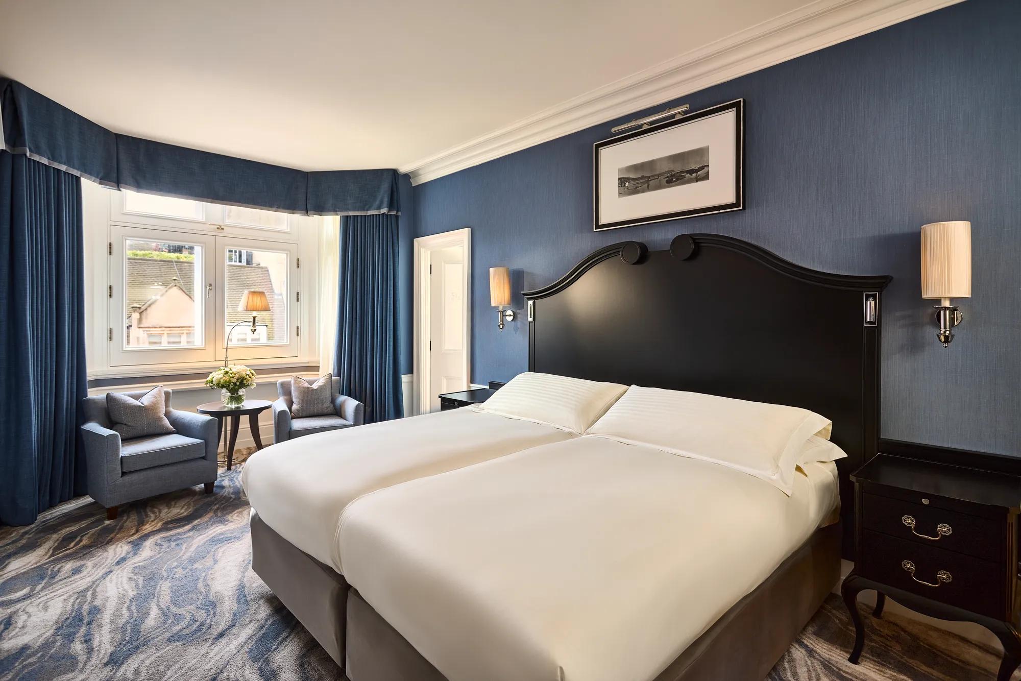 Elegant guest room with twin beds, blue walls, armchairs by window, and bedside tables.