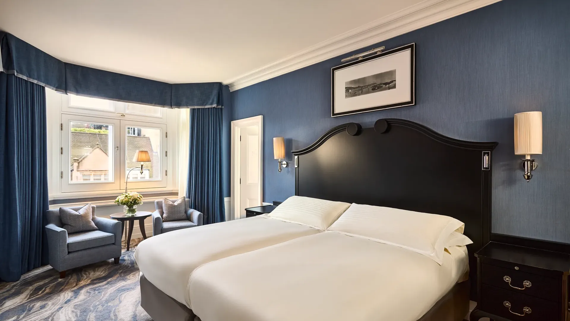 Elegant guest room with twin beds, blue walls, armchairs by window, and bedside tables.