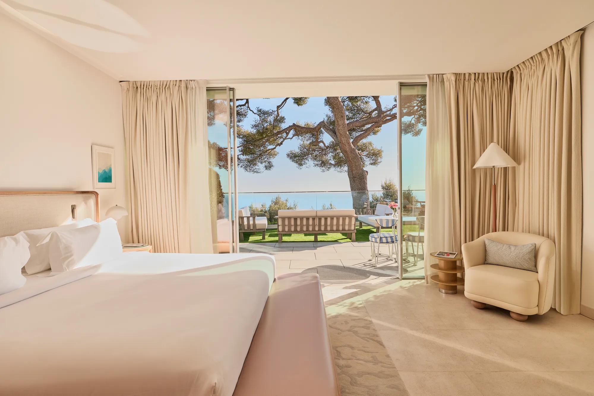 Suite d'hôtel avec lit king-size, fauteuil incurvé et lampadaire. Portes vitrées ouvertes encadrant une terrasse avec pin et vue sur la Méditerranée.