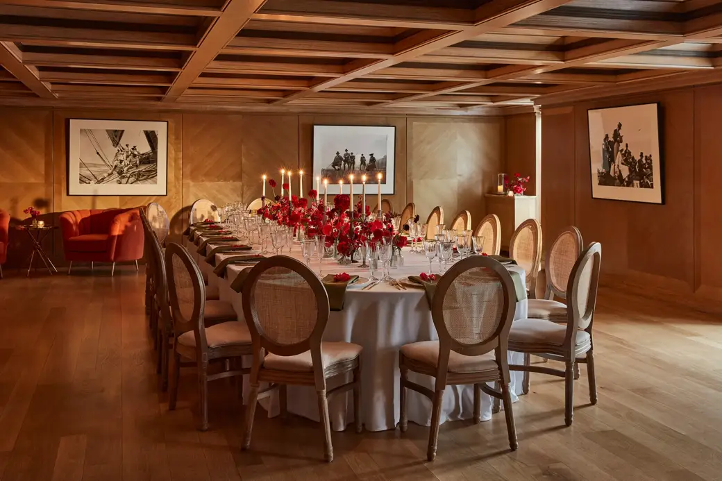 Elegant wood-panelled dining room with a long round table set with red floral centrepieces and candlelight.