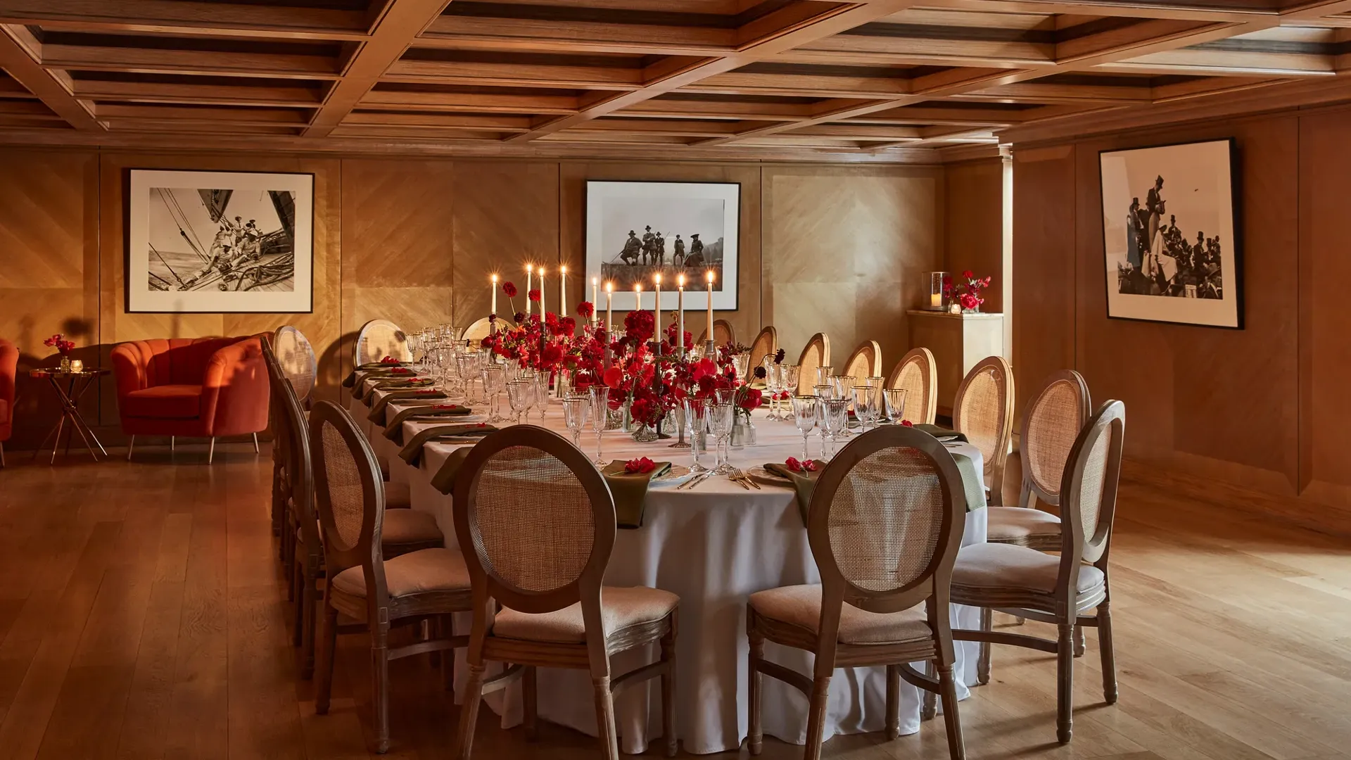 Elegant wood-panelled dining room with a long round table set with red floral centrepieces and candlelight.