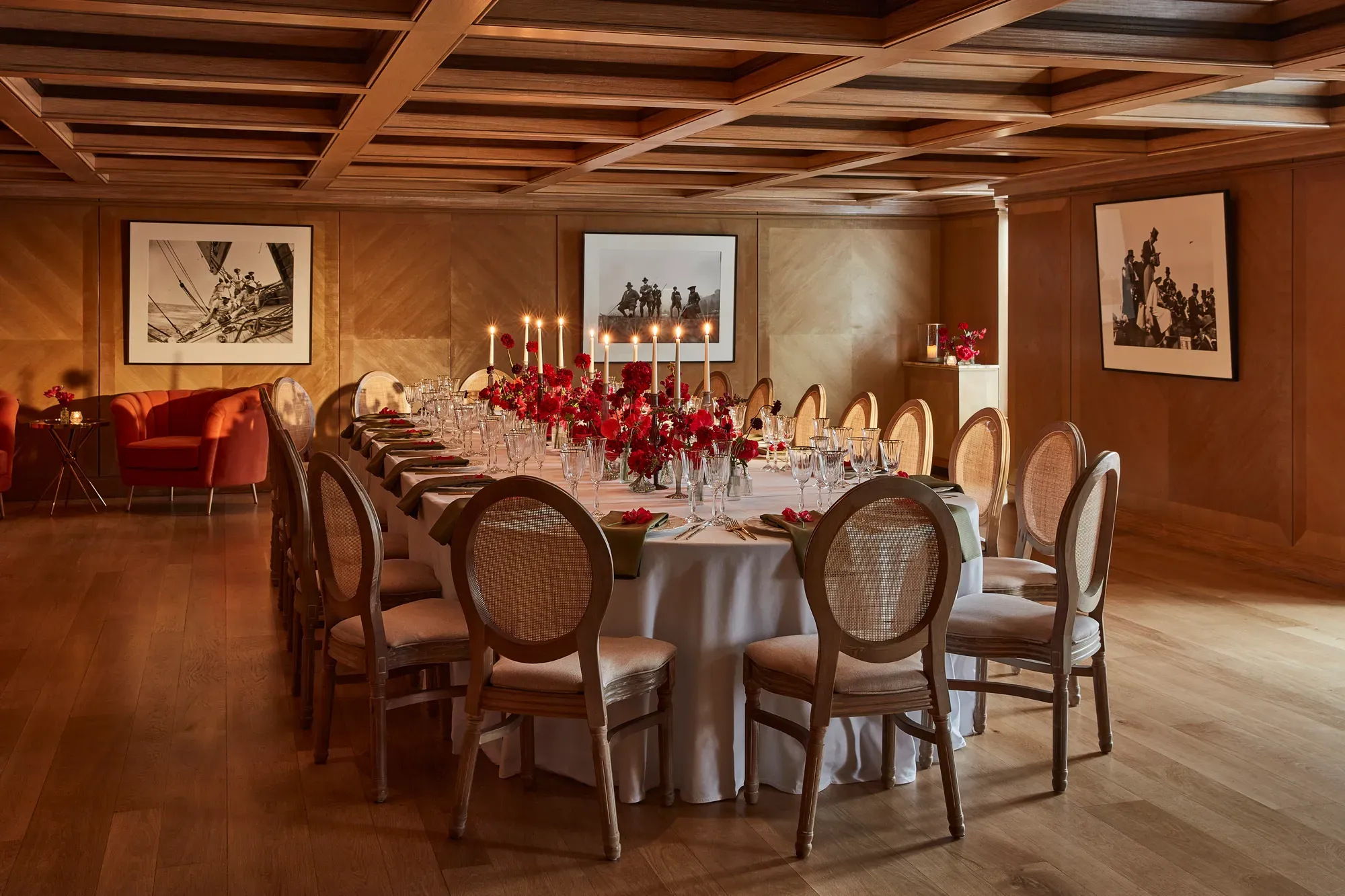 Elegant wood-panelled dining room with a long round table set with red floral centrepieces and candlelight.