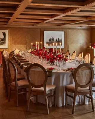 Elegant wood-panelled dining room with a long round table set with red floral centrepieces and candlelight.