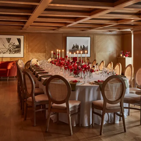 Elegant wood-panelled dining room with a long round table set with red floral centrepieces and candlelight.