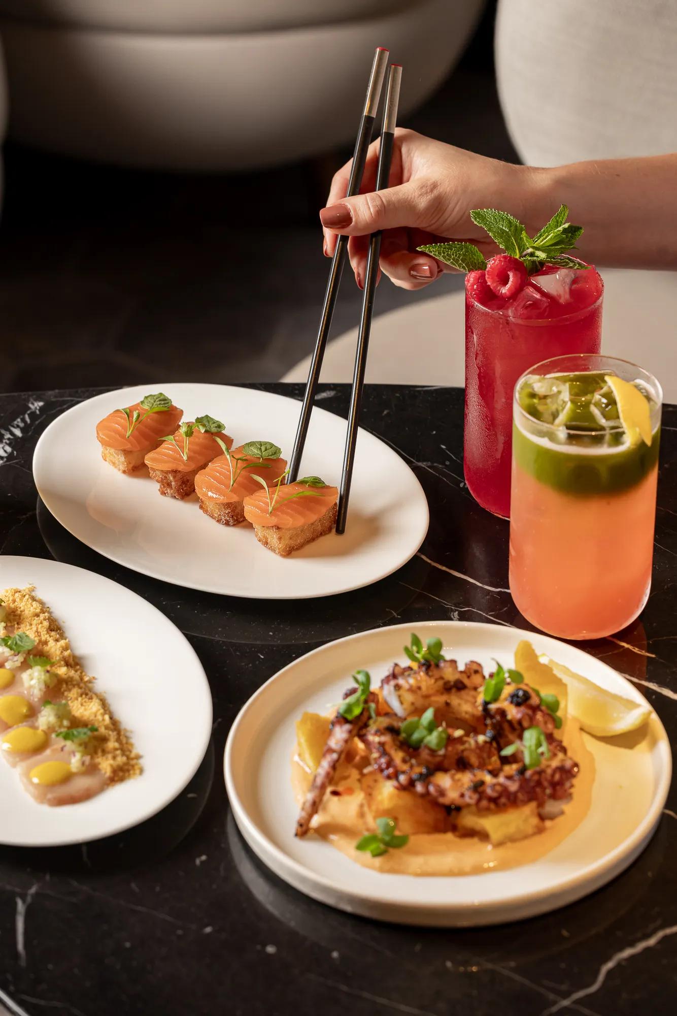 Assorted small plates with salmon bites and grilled octopus, served alongside two colourful cocktails on a dark table.
