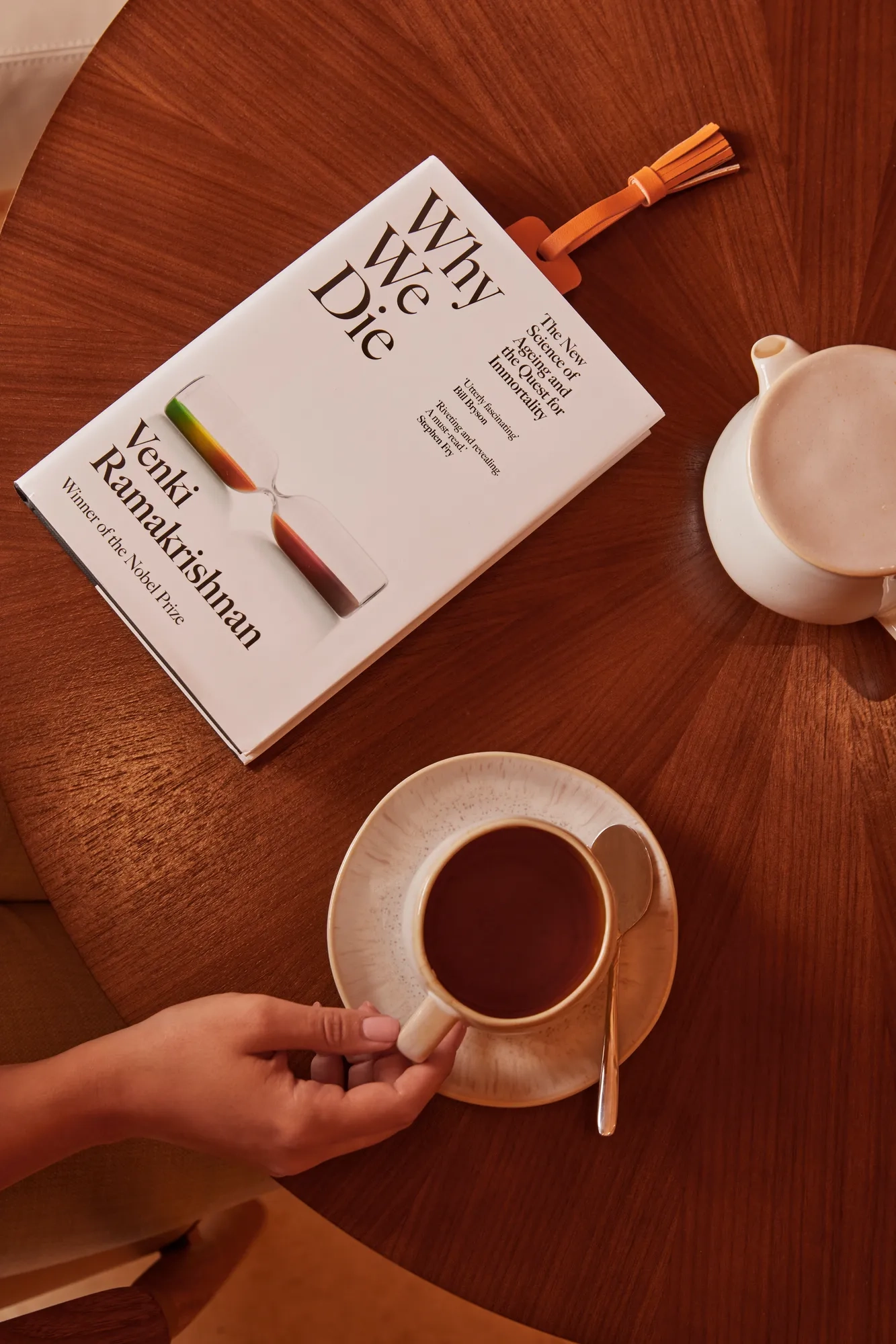 Top-down view of a coffee cup held by a hand beside the book Why We Die on a round wooden table.