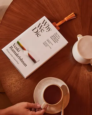 Top-down view of a coffee cup held by a hand beside the book Why We Die on a round wooden table.