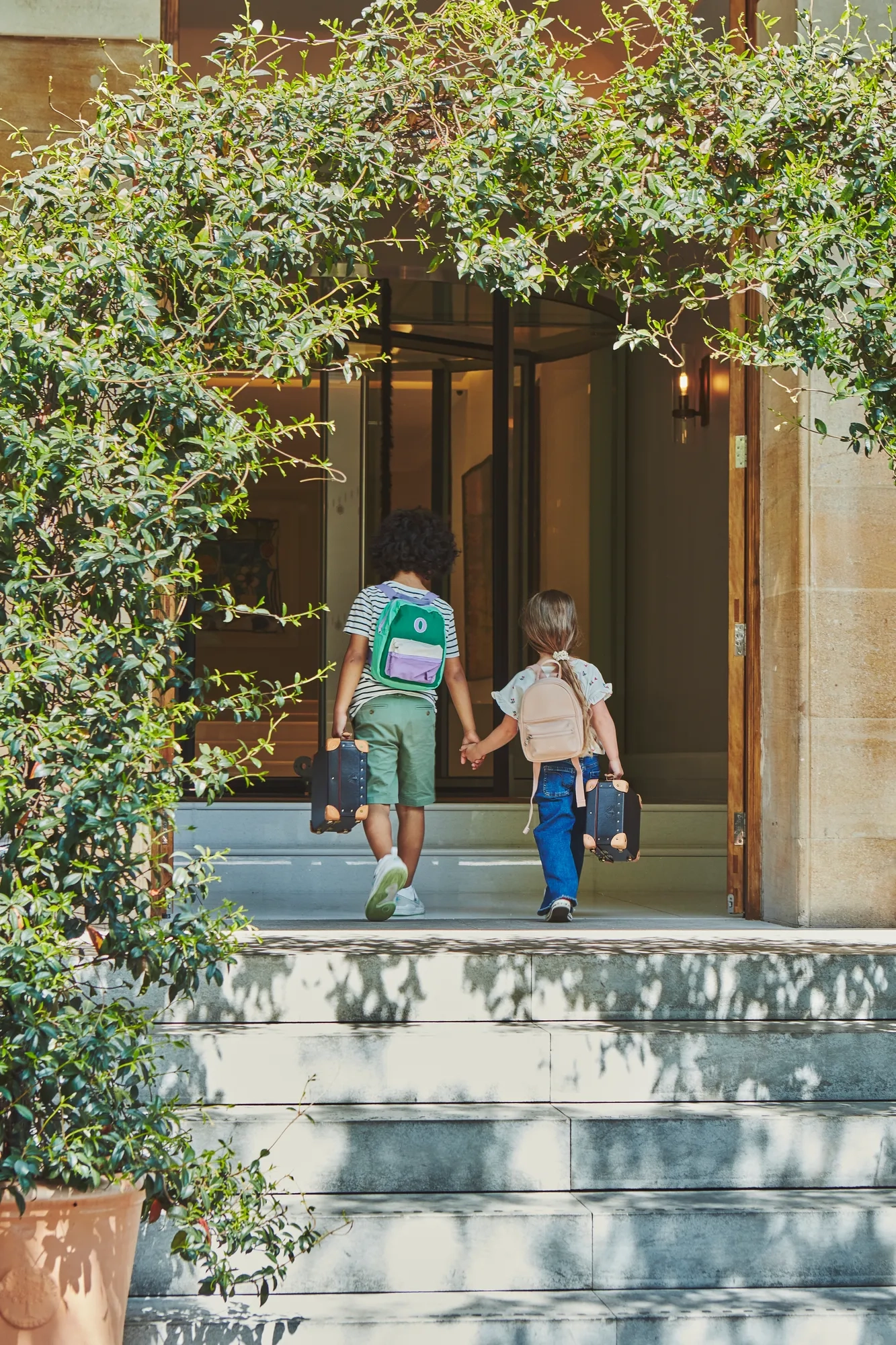 Two children holding hands walk up stone steps into a building entrance framed by lush green foliage.