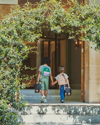 Two children holding hands walk up stone steps into a building entrance framed by lush green foliage.