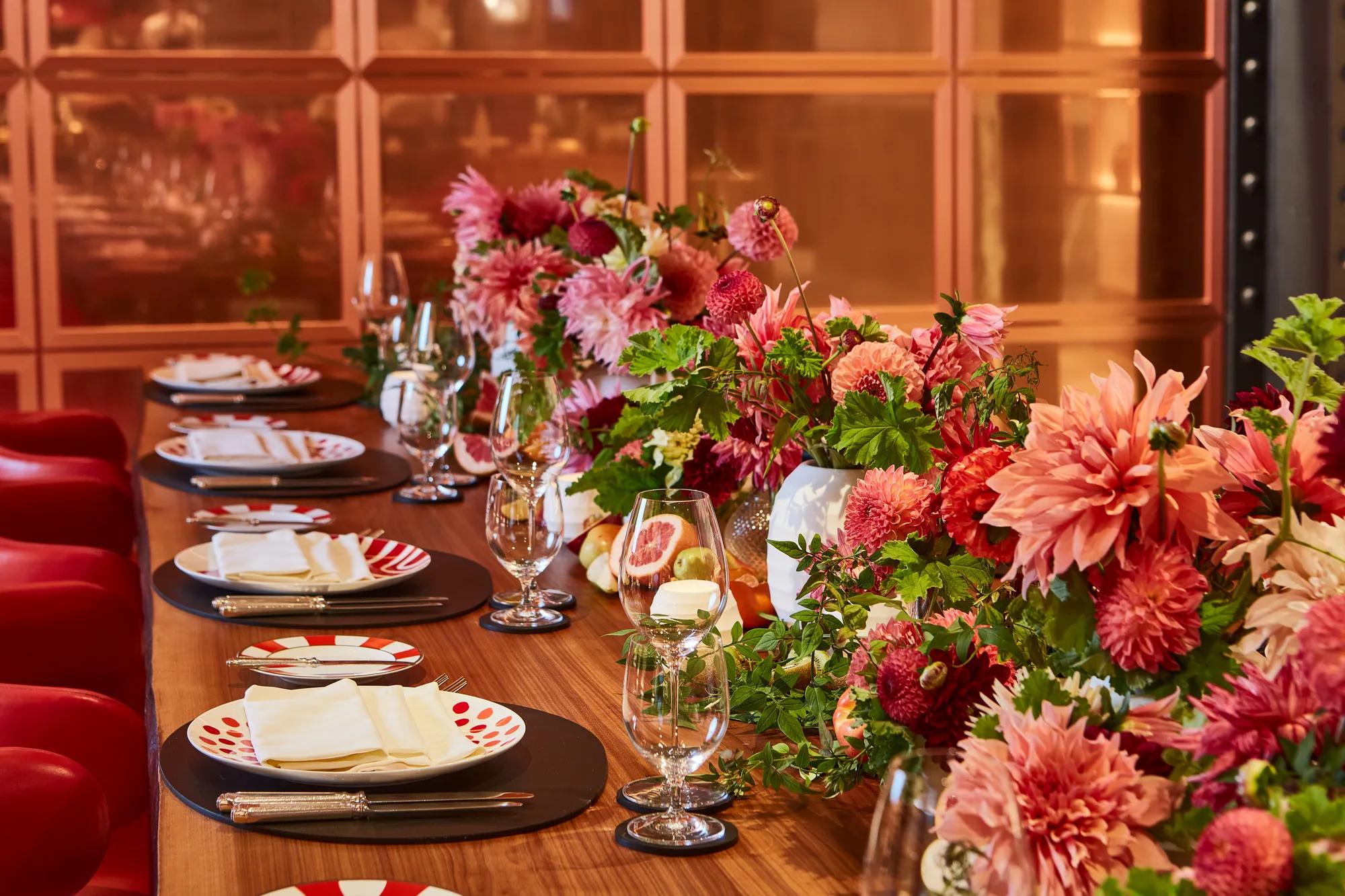 Close-up of a dining table decorated with vibrant pink and red dahlias, greenery, and fresh fruit. Each place setting includes patterned red-and-white plates, crystal glassware, and folded napkins, creating a lively and elegant atmosphere.