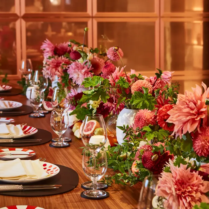 Close-up of a dining table decorated with vibrant pink and red dahlias, greenery, and fresh fruit. Each place setting includes patterned red-and-white plates, crystal glassware, and folded napkins, creating a lively and elegant atmosphere.