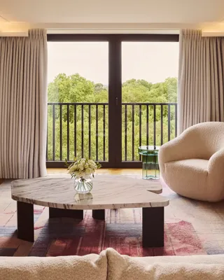 Bright living room with sofa, marble coffee table, armchair, and balcony doors overlooking trees.