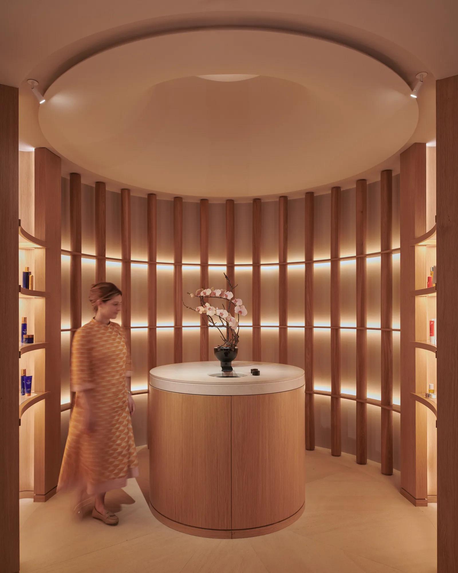A woman in a patterned dress walks through a softly lit circular alcove at The Maybourne Riviera, featuring curved wooden panels, a round table with orchids, and warm ambient lighting.