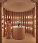 A woman in a patterned dress walks through a softly lit circular alcove at The Maybourne Riviera, featuring curved wooden panels, a round table with orchids, and warm ambient lighting.