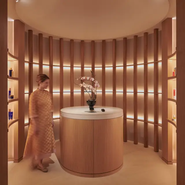 A woman in a patterned dress walks through a softly lit circular alcove at The Maybourne Riviera, featuring curved wooden panels, a round table with orchids, and warm ambient lighting.