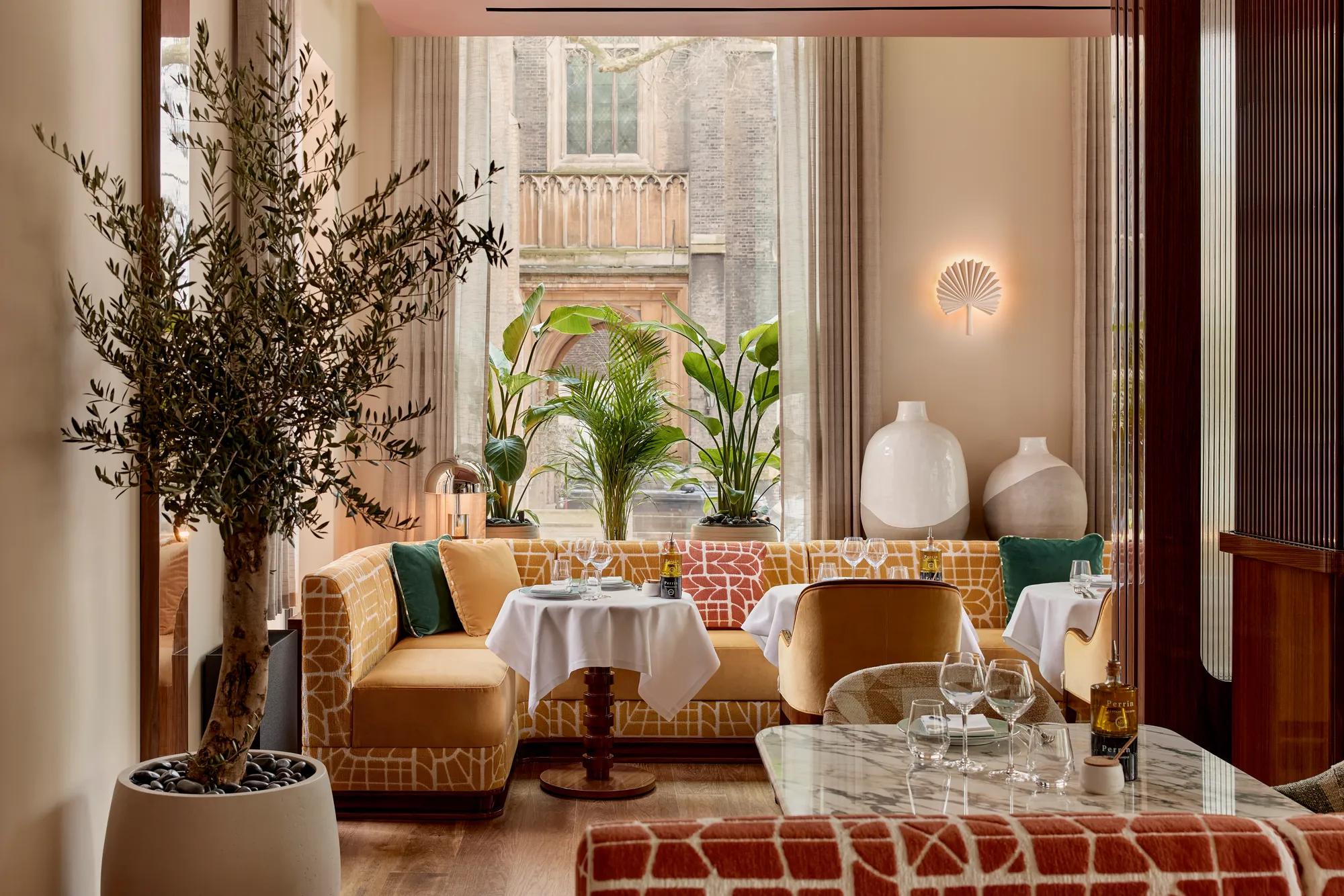 Restaurant interior with patterned seating, marble tables, and large windows framed by plants, creating a bright, inviting space
