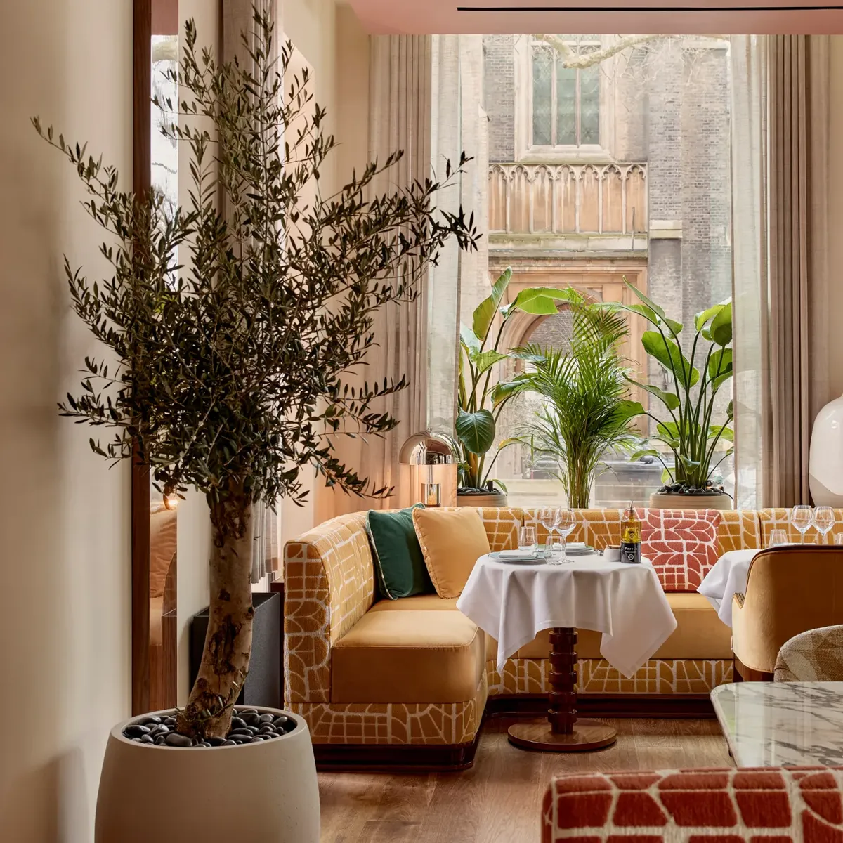 Restaurant interior with patterned seating, marble tables, and large windows framed by plants, creating a bright, inviting space
