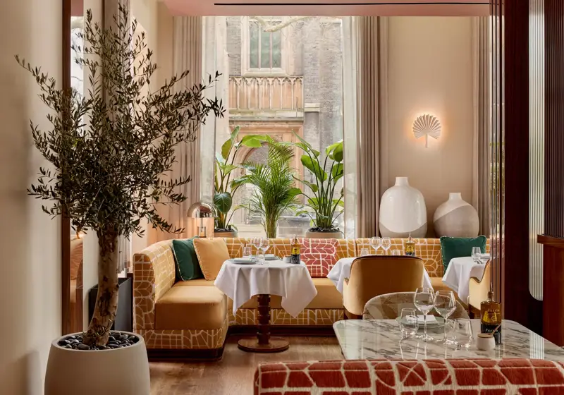 Restaurant interior with patterned seating, marble tables, and large windows framed by plants, creating a bright, inviting space