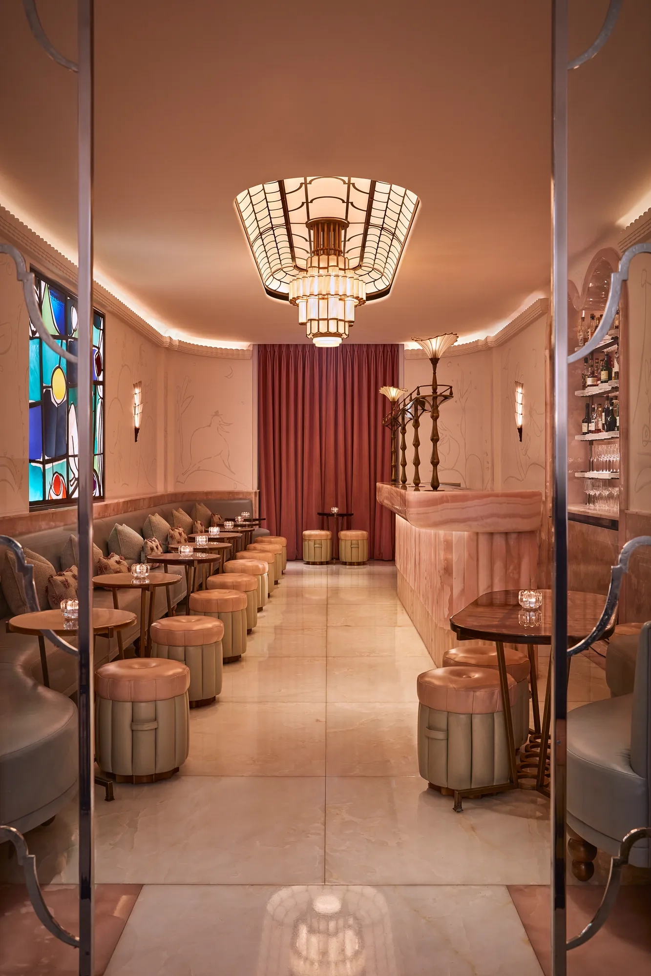 Art Deco–inspired bar with blush pink marble counter, upholstered stools, and soft lighting from a geometric chandelier. A stained-glass window and deep red curtains add warmth and elegance to the intimate space.